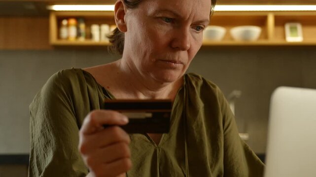 Woman holding credit card with concerned expression while working on laptop in home office, 4K