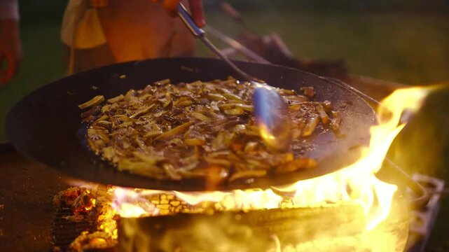 Chef stirring sliced potatoes on large flat griddle over crackling outdoor fire.