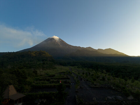 Aerial view of Bunker Kaliadem Merapi and the smoking peak of Mount Merapi volcano surrounded by lush green forests and a winding path Kecamatan Pakem, Daerah Istimewa Yogya, Indonesia.