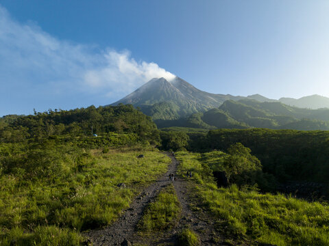 Aerial view of Bunker Kaliadem Merapi and the smoking crater of Mount Merapi volcano with lush green hills and a dirt path under a blue sky Kecamatan Pakem, Daerah Istimewa Yogya, Indonesia.