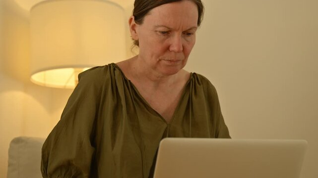 Mature woman typing on laptop in living room workspace, concentrated remote work moment with warm indoor lighting. 4K.