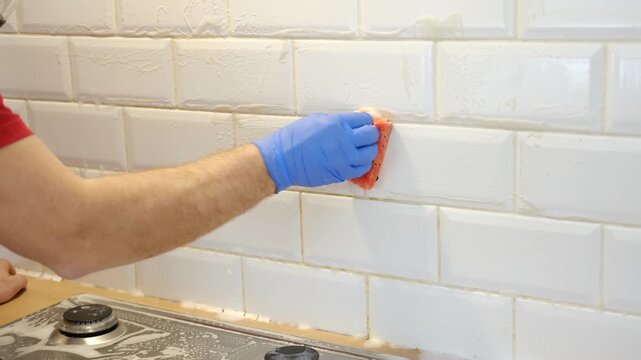 Thoroughly cleaning the grout between tiles. Manually cleaning stains on white tiles. Close-up of a gloved hand cleaning white tiles. A variety of tile cleaning products. The problem of cleaning.