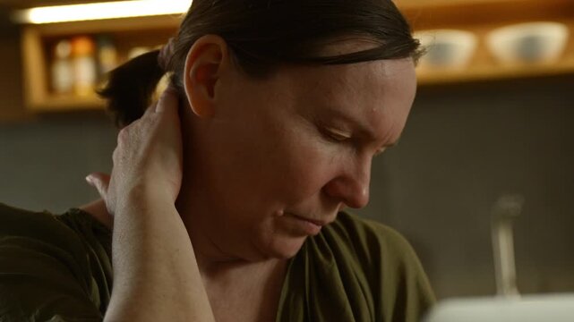 Mature woman with eyes closed rubbing back of neck in discomfort near laptop, fatigue and muscle tension from computer work. 4K.