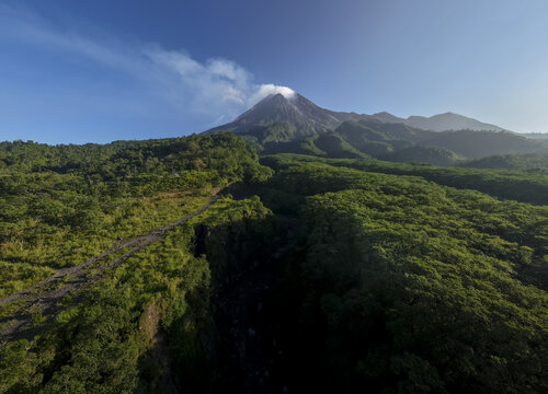 Aerial view of Bunker Kaliadem Merapi and the smoking peak of Mount Merapi volcano surrounded by lush green forests and rolling hills Kecamatan Pakem, Daerah Istimewa Yogya, Indonesia.