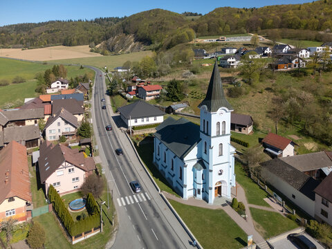 Aerial view of the light blue church of Saint Joseph with its tall spire, surrounding village houses, and a road winding through green hills Kriva, Zilina Region, Slovakia.