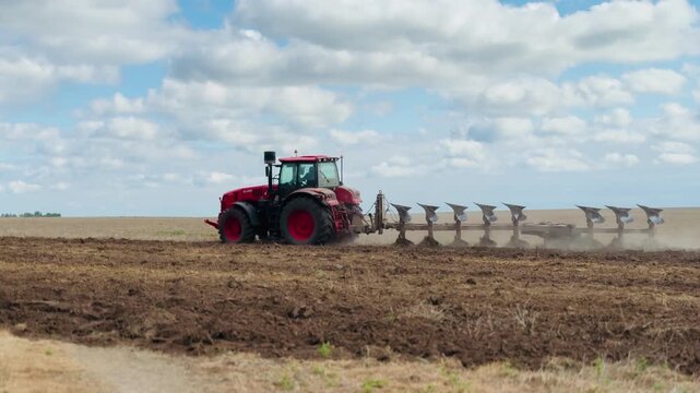 Farm tractor pulling a metal plow through dry field soil under cloudy daytime sky for agricultural work and cultivation footage
