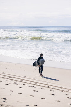Surfer walking on beach carrying surfboard looking at ocean