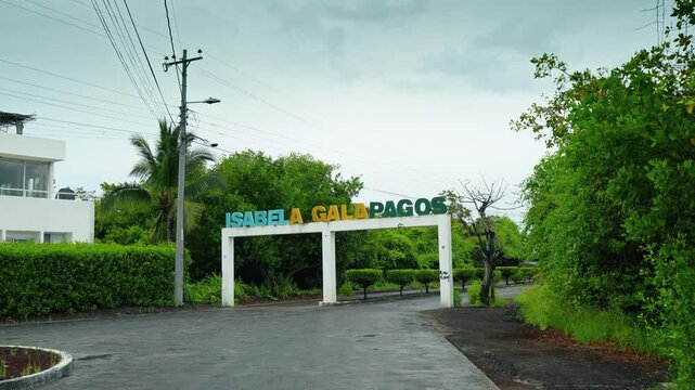 Approaching the arch on the road with the sign Isabela Galapagos. White building and lush greenery around.