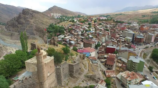 Aerial ancient hilltop fortress overlooking dense town houses in rugged valley under soft daylight. Birds eye shot stone castle tower crumbling ramparts and crowded rooftops beside river.