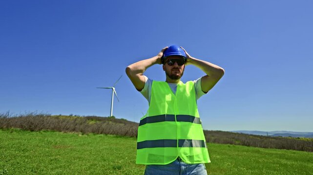 A windmill engineer is in a green field wearing a safety vest and sunglasses. He holds a blue hard hat and looks towards the wind turbines in the distance during bright sunny weather.