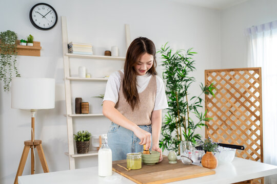 Asian young woman preparing matcha by stirring green tea powder in bowl.