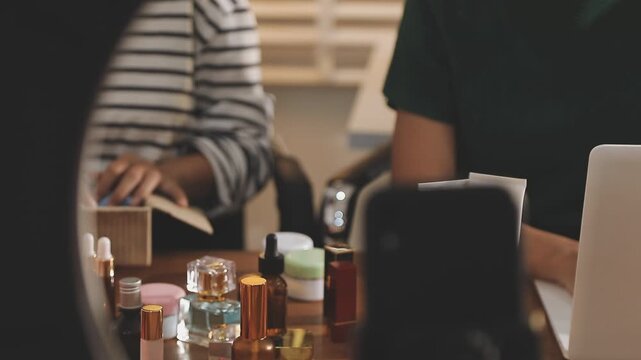 Young beautiful Asian girl reviewing cosmetic and skin care product with new collection's promotion selling on social media online live blurred phone screen surrounded beauty merchandise. Stratagem.