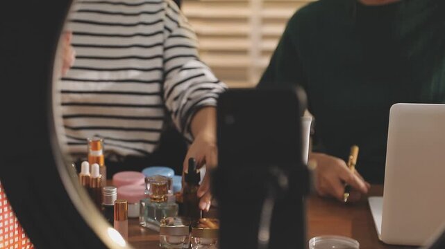 Young beautiful Asian girl reviewing cosmetic and skin care product with new collection's promotion selling on social media online live blurred phone screen surrounded beauty merchandise. Stratagem.