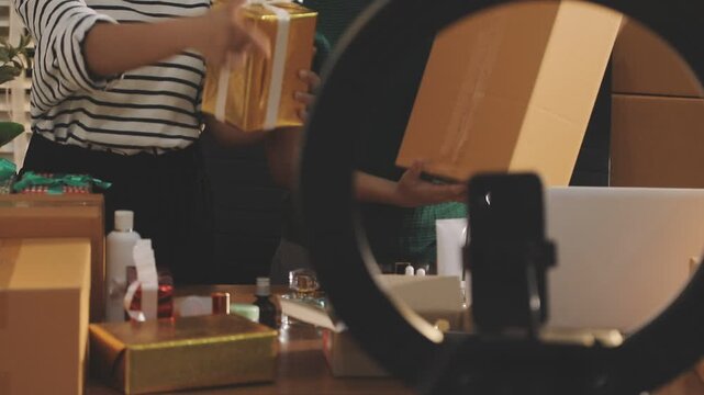 Young beautiful Asian girl reviewing cosmetic and skin care product with new collection's promotion selling on social media online live blurred phone screen surrounded beauty merchandise. Stratagem.