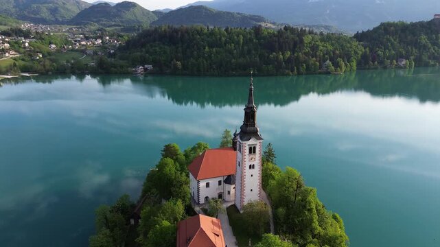 Cinematic aerial orbit shot of the Pilgrimage Church of the Assumption of Mary on the island of Lake Bled, Slovenia. Crystal clear turquoise water and lush green forest in summer
