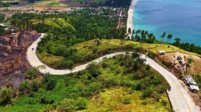 A winding road snakes along a lush green hillside overlooking the turquoise sea at San Vicente viewpoint in the Philippines, captured from a drone