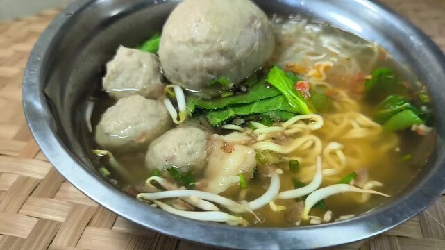 Steaming noodle bowl with meatballs and greens. Metal bowl filled with warm, flavorful broth. Noodles float beneath tender meatballs. Bean sprouts and herbs add freshness. This image exudes a sense of