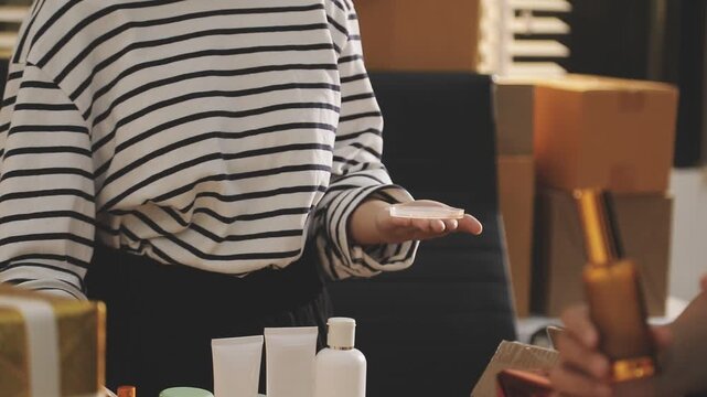 Young beautiful Asian girl reviewing cosmetic and skin care product with new collection's promotion selling on social media online live blurred phone screen surrounded beauty merchandise. Stratagem.