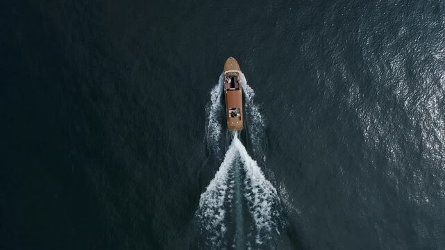 Aerial drone view of wooden boat moving across Lake Como with white wake trailing behind on deep blue water surface with minimal composition and smooth texture