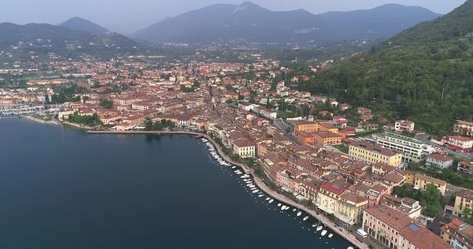 Aerial drone view of Salo town with dense terracotta rooftops along curved Lake Garda shoreline with calm water and surrounding green hills under soft daylight