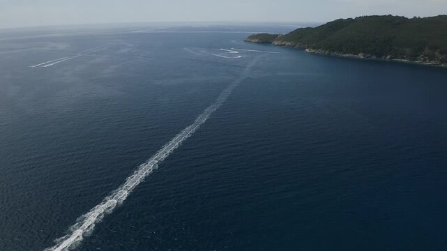 Aerial drone view of speedboat moving across deep blue sea leaving long straight wake trail with distant coastline near Himare under clear sky and calm water