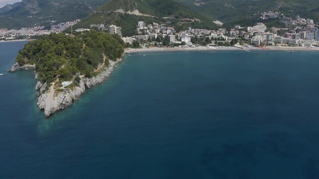 Aerial drone view of Becici Beach and Zavala Peninsula with clear blue sea curved coastline and resort buildings along shore in Budva under bright daylight