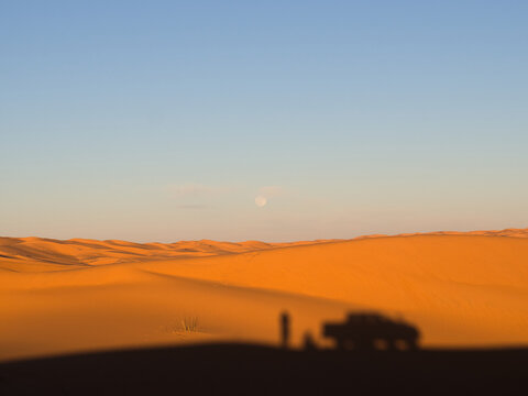 Full moon rising over glowing orange Sahara sand dunes at sunset with silhouette of person and 4x4 vehicle, Algeria