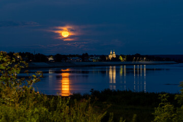 Fototapeta premium A beautiful night landscape with a full moon rising over a river. Moonlit path on the water. The Church of St. Simeon the Stylite is visible in the distance. Veliky Ustyug, Vologda Oblast, Russia.