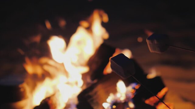 People roasting marshmallows on sticks over a crackling campfire at night, creating a warm and inviting atmosphere for a delightful treat.