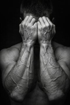 A muscular man hides his face with veiny hands in dramatic black-and-white lighting, creating a raw and emotional atmosphere on a dark background.