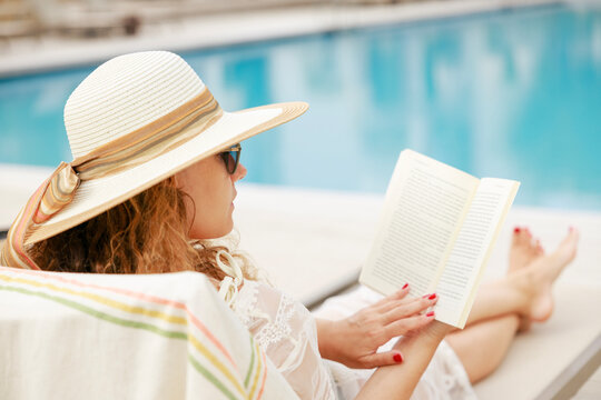 Woman reading book by poolside relaxing during summer vacation
