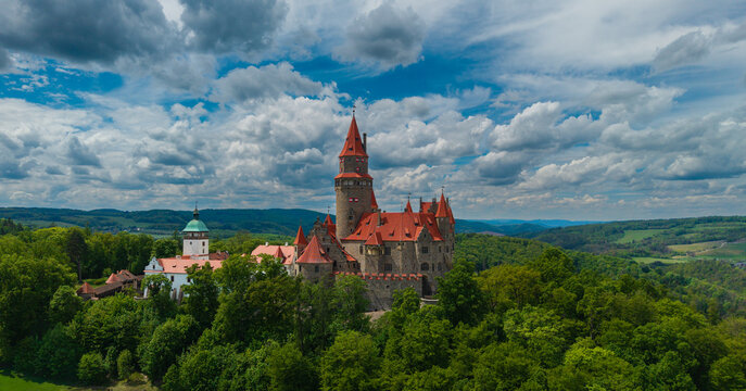 Aerial view of Bouzov Castle with its iconic red-roofed towers and stone walls nestled among lush green forests under a dramatic cloudy sky Bouzov, Olomoucky kraj, Czechia.