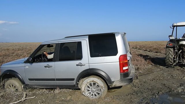 Mud stuck suv being pulled by tractor on rural field track during off road vehicle recovery. Bogged utility car rescued with tow rope across farmland trail as countryside transport gets freed.