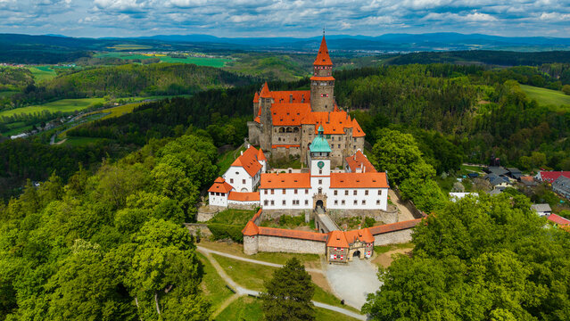 Aerial view of Bouzov Castle with its iconic red-tiled roofs and tall stone tower nestled among lush green forests and rolling hills in Bouzov, Olomoucky kraj, Czechia.