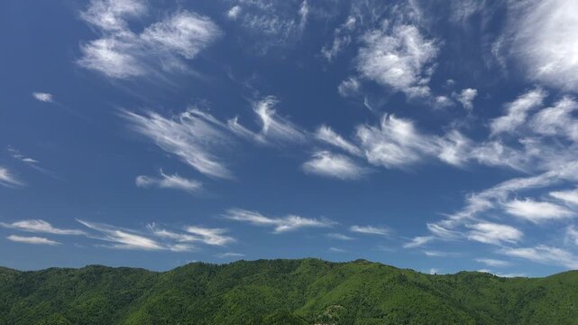 Wispy cirrus stretch above alpine forest and green hills, opening toward distant mountain ridges. Bright high clouds and quiet woodland create a tranquil upland view with clear atmosphere.