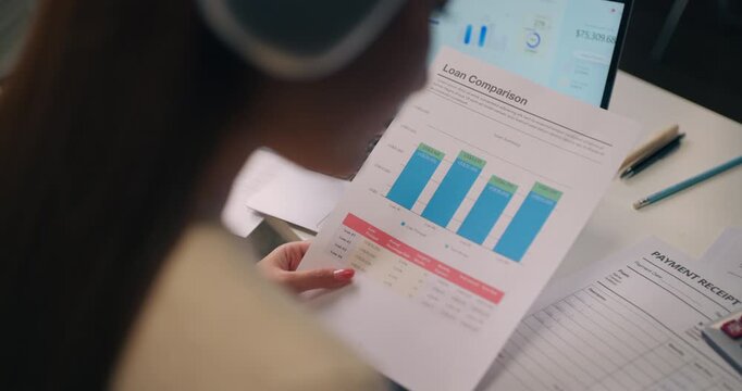 Close-up of female holding loan comparison paper with bar graph and calculator on desk. Mortgage planning, debt analysis, and financial investment research.