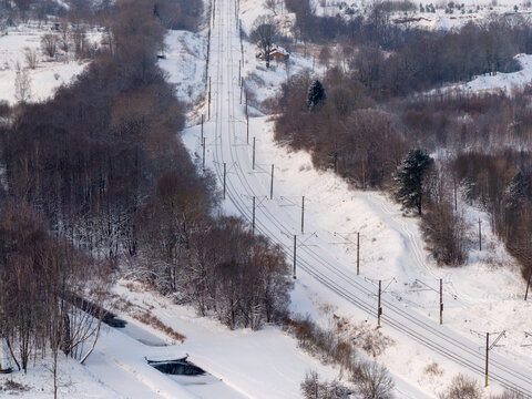 Aerial view of electrified railway tracks cutting through a dense snow-covered forest and rolling hills in the winter countryside Lithuania.