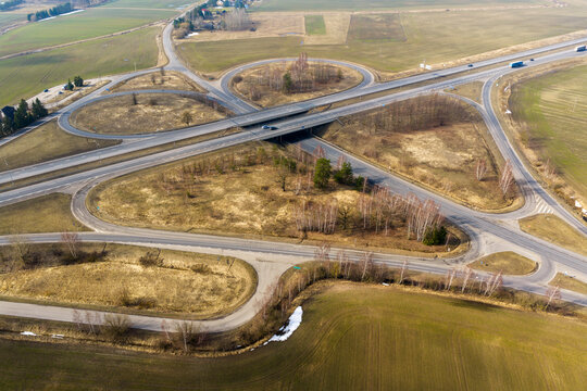 Aerial view of a highway interchange and cloverleaf junction surrounded by agricultural fields and sparse trees with patches of snow in Lithuania.