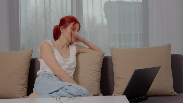 A woman relaxes on the couch, intently watching something on her laptop in a warm and inviting living room setting during the day.