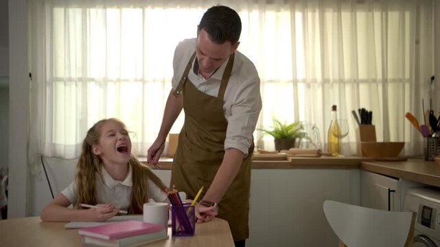 Caring father supervising his daughter while she studies and writes homework at home in a warm kitchen environment