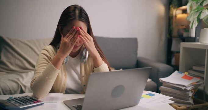 Stressed Asian female entrepreneur with headache during work at home. Burnout, overwork, financial stress, and deadline pressure.