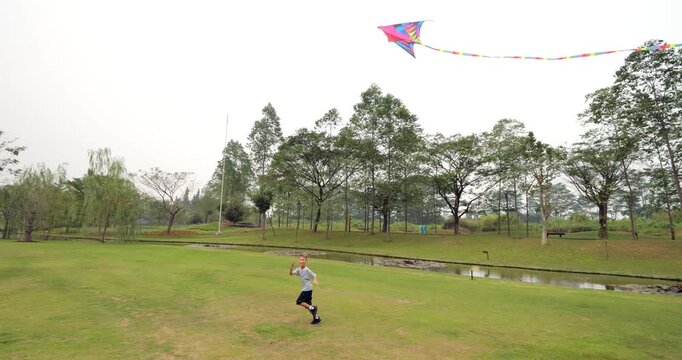 Slow-motion tracking shot of teenage boy running across lawn to generate lift for his multi-colored kite during low wind day. By towing kite and creating apparent wind through his own movement