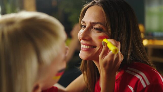 Two friends get ready for a sports event. One friend paints the other's face in team colors. Laughter fills the room as they enjoy their time together.