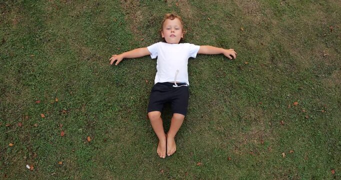 Top-down shot of young toddler boy lying on his back on short lawn with his arms spread wide. With his knees partially bent, he rubs his bare feet against grass, exploring tactile sensations