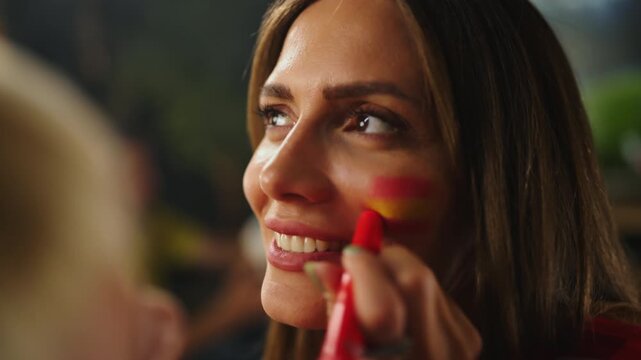 Close up of female football supporter getting national flag face paint from friend while preparing to watch sports match and cheer for favorite team.