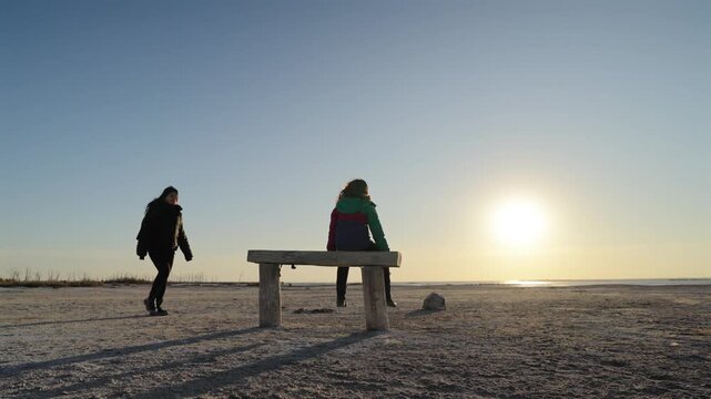 Two people meeting on bench at shoreline with one sitting and receiving an embrace under low sun creating long shadows and warm light suggesting support connection and affection static shot