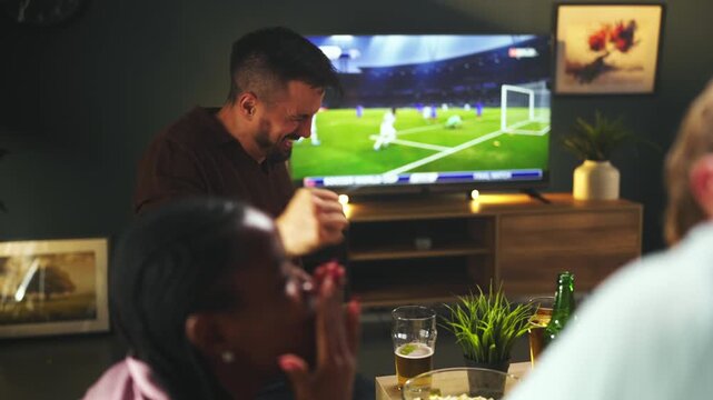 Excited sports fans celebrating goal and cheering together while watching football or soccer match on television in cozy home atmosphere.