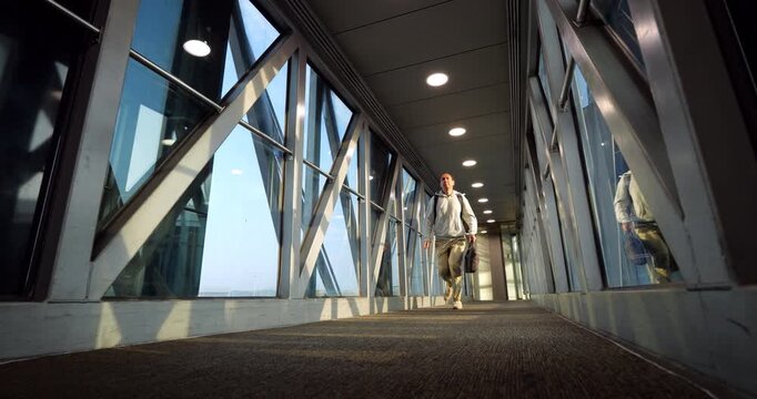 Digital nomad traveler running through passenger boarding bridge, rushing from airliner to catch connecting flight, low angle, slow motion shot. Male traveler appears in hurry