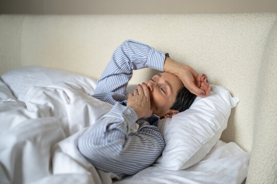 Dreamy senior woman comfortably lying in bed covered in cozy blanket and yawning after waking up in morning, sleepy lazy elderly female awakening at early hours after good night rest and sweet dream.
