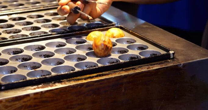 Close-up view of street food vendor turning last few hot takoyaki balls with metal sticks on cast-iron pan at Jalan Alor in Bukit Bintang. Sizzling snack is nearly ready to serve
